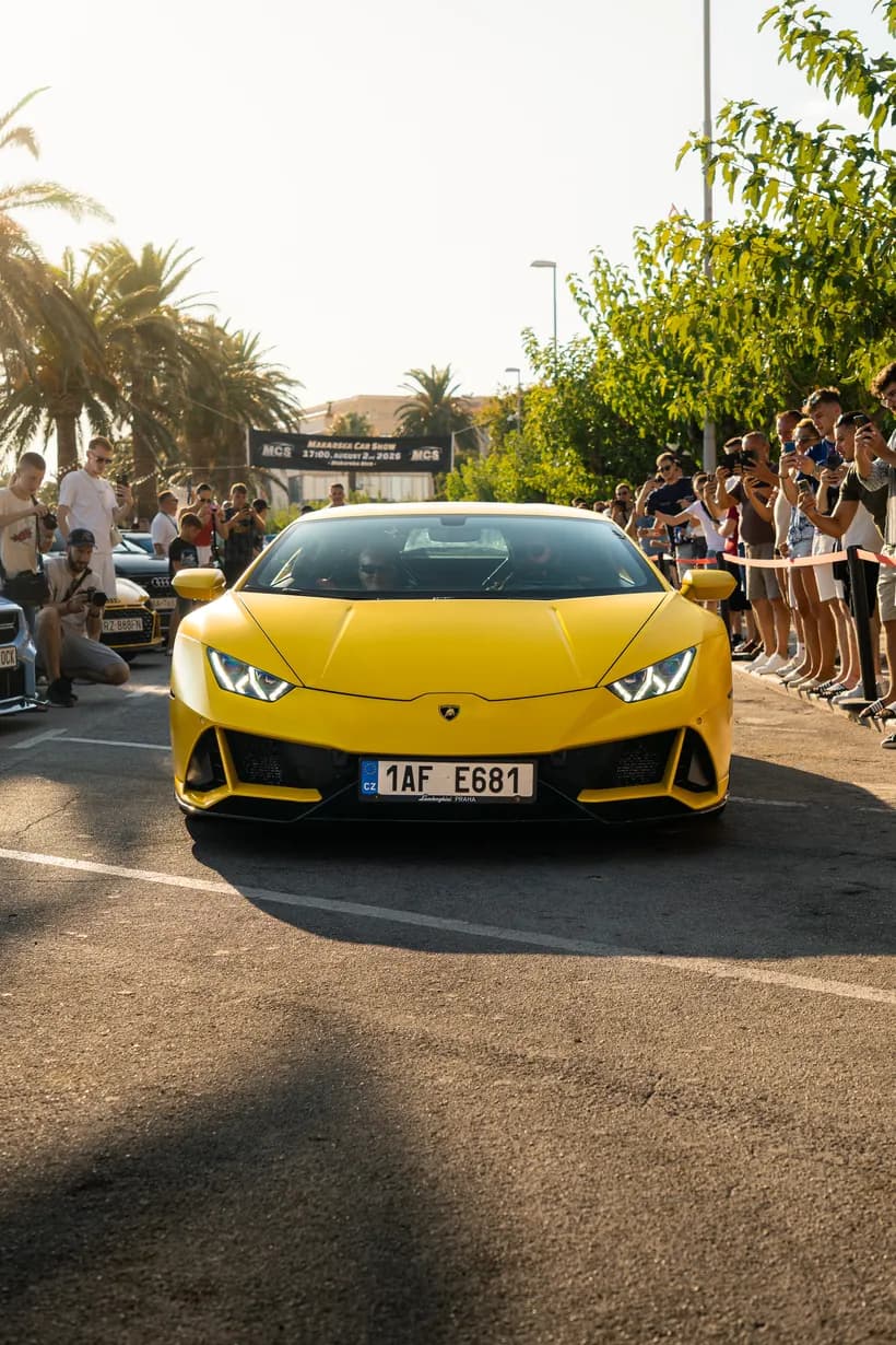 A yellow Lamborghini Huracan seen from the front left at a car meet, crowd behind a barrier, palm trees overhead.