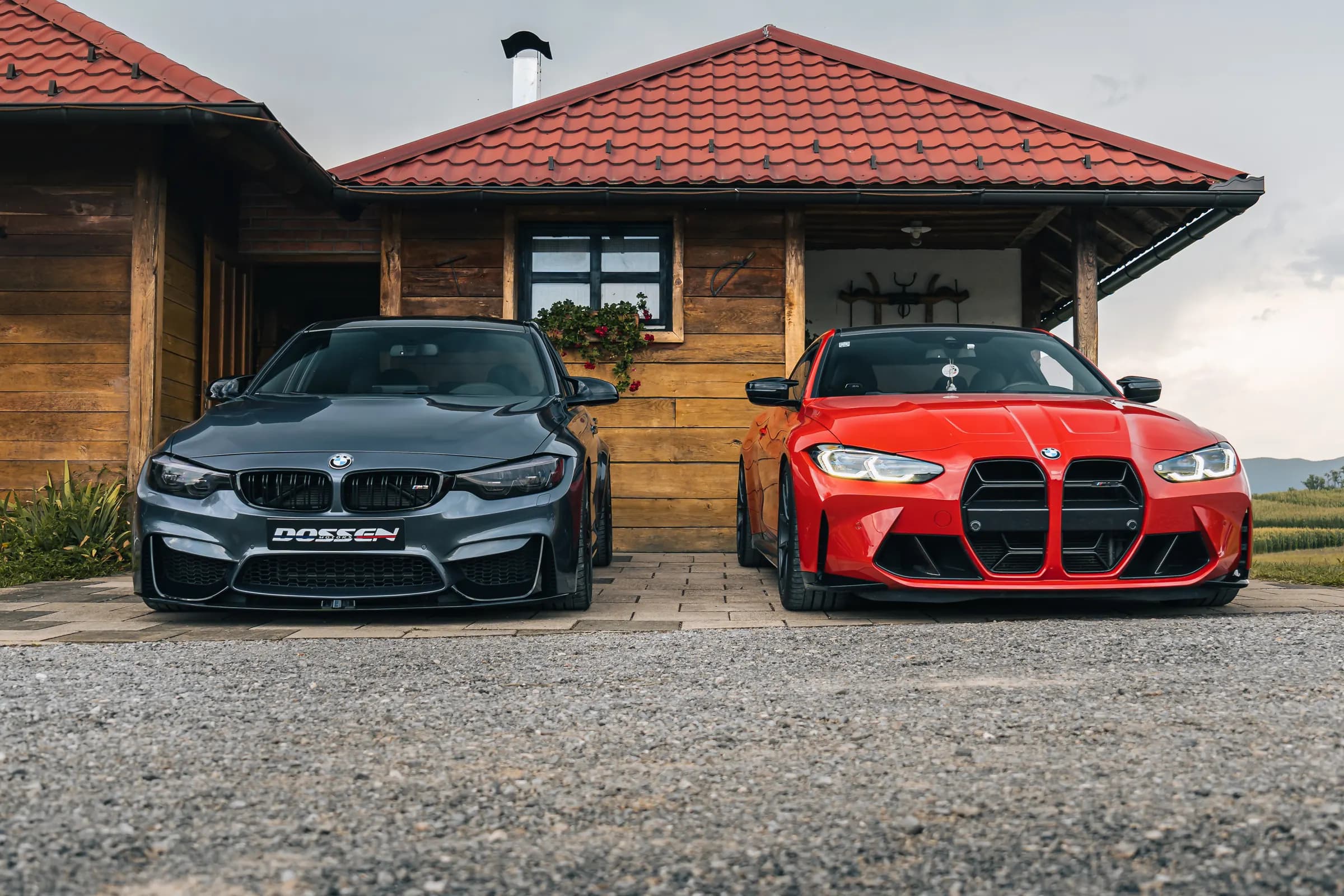 Two BMW M4s parked side by side on gravel in front of a wooden house at a car meet, one matte grey and one red, photographed head on in flat morning light.